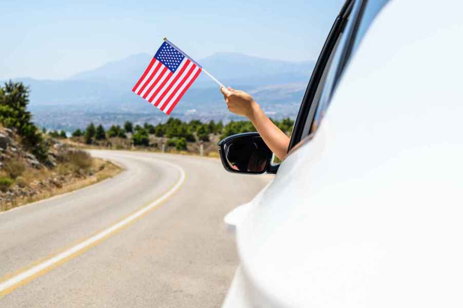 Hand holding small American flag out of white car driving on the highway in close side view