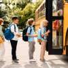 Row of students wait to board a bright yellow school bus, trees visible in the background.