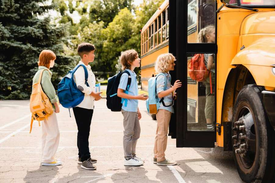 Row of students wait to board a bright yellow school bus, trees visible in the background.