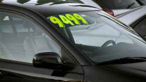 A black used car with price sticker parked at a used car dealer in close view of windshield right front angle