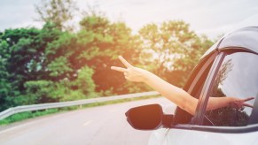 The bare arm of a woman flashing a peace sign outside the window of her white sedan on the highway.