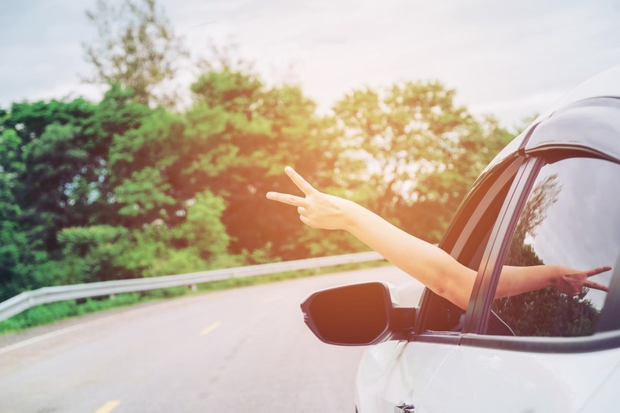 The bare arm of a woman flashing a peace sign outside the window of her white sedan on the highway.