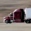 A semi-truck with dark purple cab and white trailer driving fast on highway in left profile view