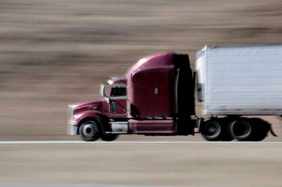 A semi-truck with dark purple cab and white trailer driving fast on highway in left profile view